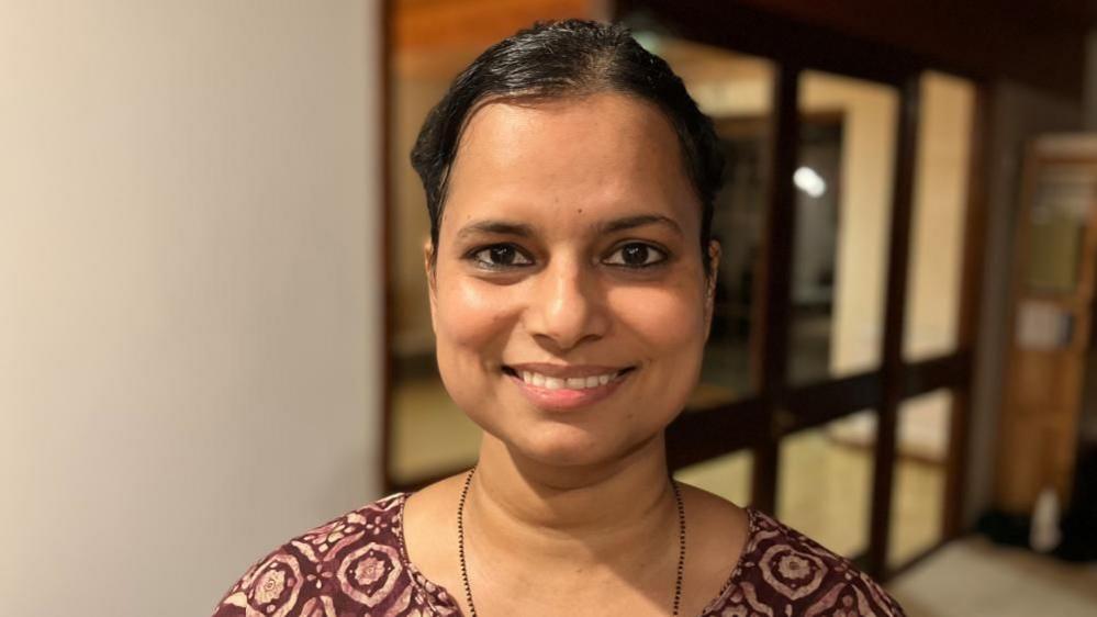 Ashwini Mahatre looking at the camera. She is smiling, with her dark hair tied back. She has a necklace and a burgundy and cream outfit. 