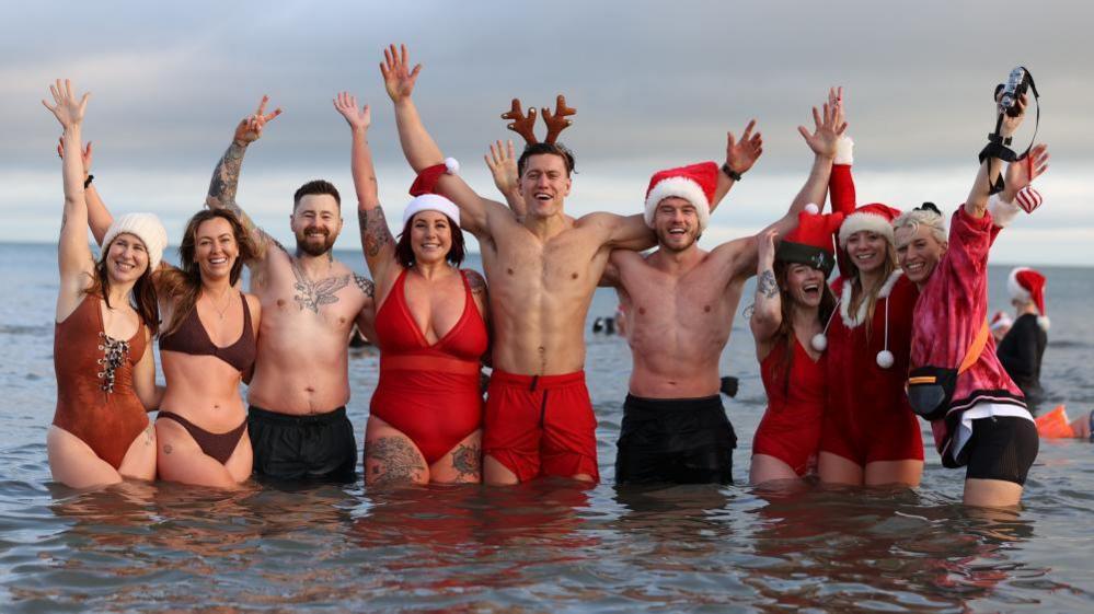 A group of people in the sea wearing red and black swimming costumes, Santa hats and antlers. They are smiling at the camera and have arms in the air.