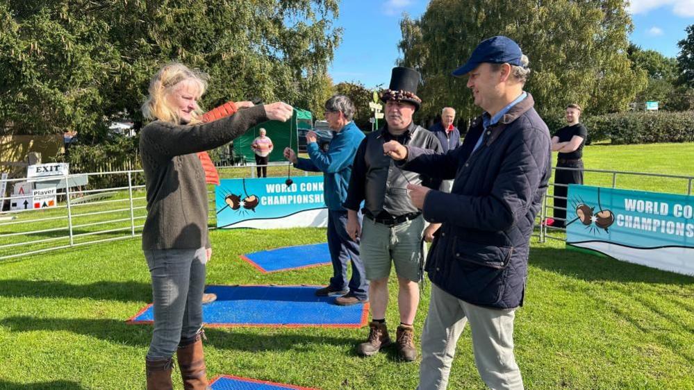 A woman with blond hair and a man in a blue cap play conkers while a man in a large black hat watches. Another game is going on behind them. There are blue mats on the floor and "World Conker Championship" signs around a fenced-off arena.