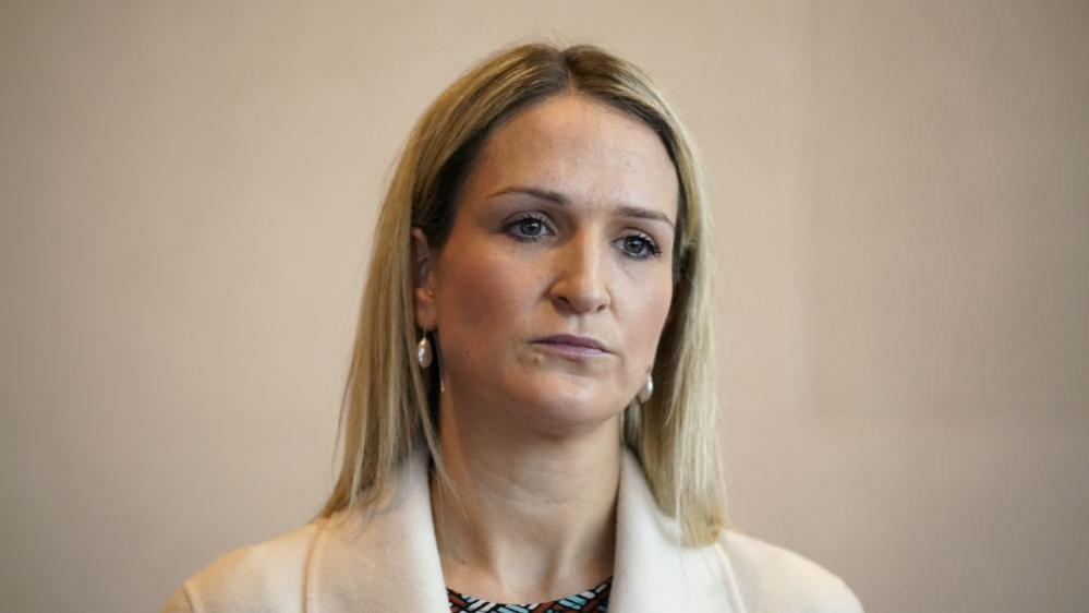 A woman with long blonde hair wearing silver earrings and a cream blazer.