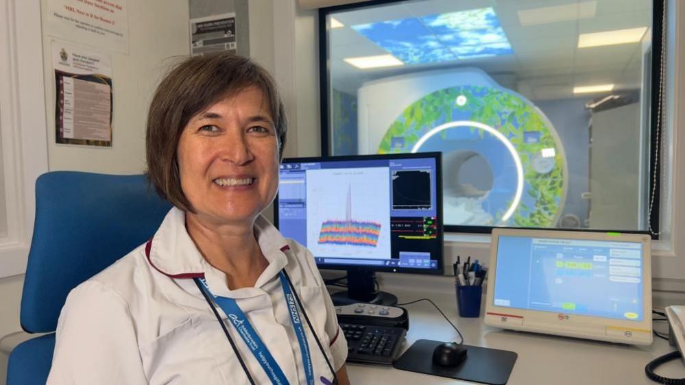 Ilse Patterson, wearing a clinical uniform, sitting on a chair in front of a MRI scanner and monitors