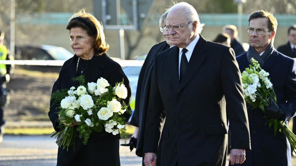 Queen Silvia and King Carl XVI Gustaf walk side-by-side, wearing all black, with sombre expression. Queen Silvia carries a bouquet of white flowers.