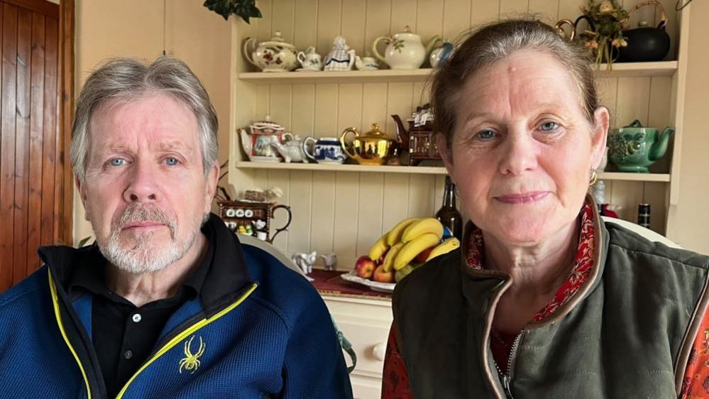 David and Tracey sat side by side; he's wearing a royal blue fleece, and she's wearing an orange patterned top with a khaki gilet over the top. They're sat in front of a cabinet with tea pots, mugs, and a fruit bowl. 