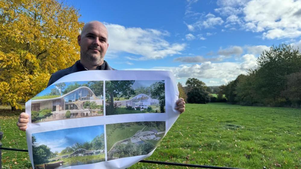 Ashley Broughton has a shaved head and close-cropped beard, and stands in the area where the new house will be built, which is laid to grass with trees bordering the site. He is wearing a grey zipped jacket, with black shirt under. He is holding a print of artists impressions of the property he designed.