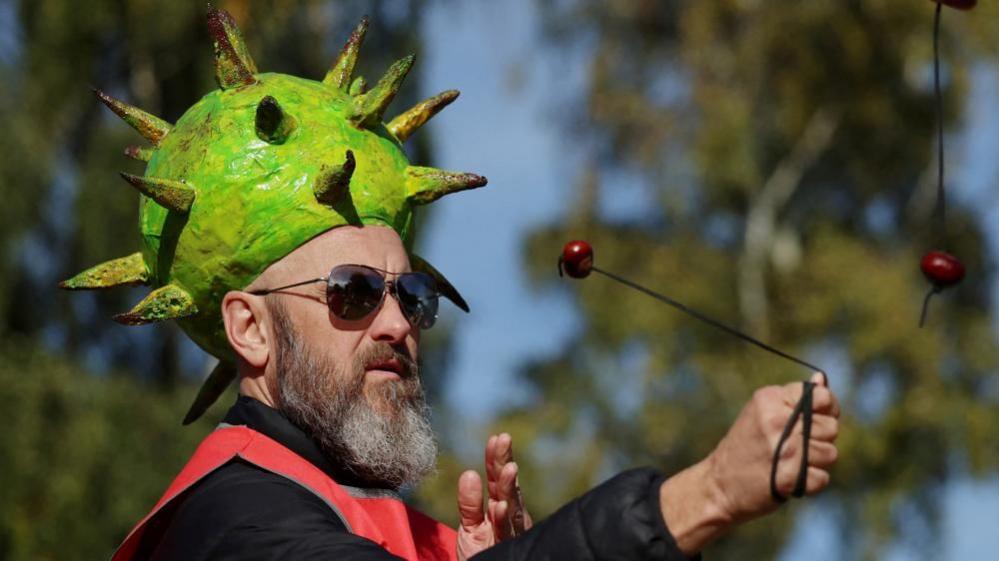 A man with a greying heard wearing sunglasses and a hat shaped like a green conker shell