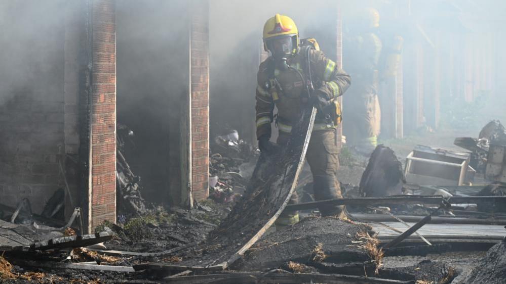 A firefighter in full breathing apparatus and a yellow helmet holds a piece of metal sheeting, which is in the middle of debris outside a row of burned-out garages. Another firefighter is standing in the entrance to another garage just behind him. There is a haze of smoke in the air around them.