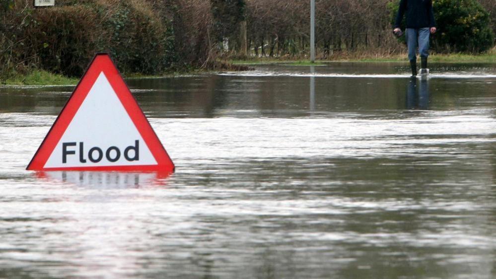 An area flooded with water and a red, triangular warning sign that reads "flood" is covered with flood-water at the bottom. A man's legs and torso can be seen at the top right of the image, he is walking through the ankle-deep water. There are also trees and bushes in the background that are not underwater. 