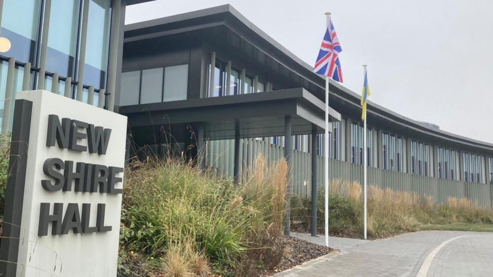 A sign saying "New Shire Hall" next to the modern headquarters of Cambridgeshire County Council in Alconbury.