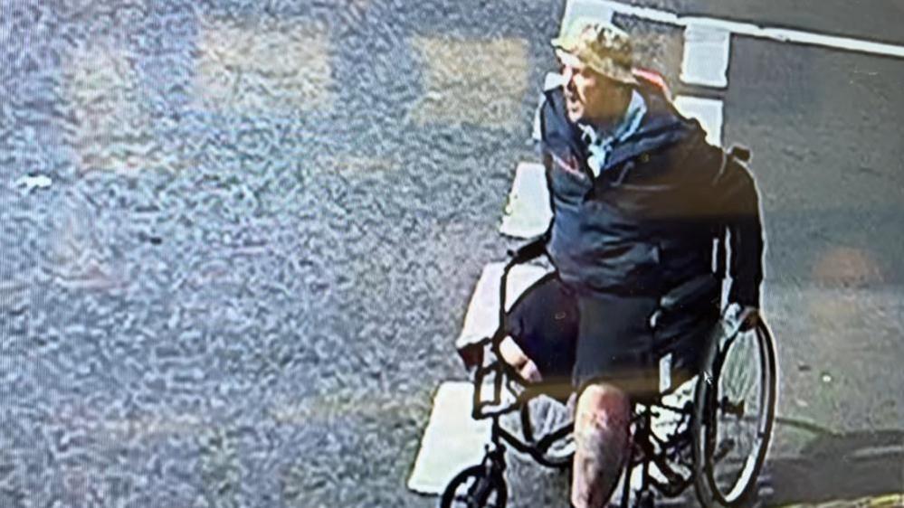 Michael McQueen in his wheelchair in the junction of St Peter's Road to King Street in Great Yarmouth. McQueen is wearing black shorts, and this shows he has had his right leg amputated at the knee. He is wearing a blue jacket and light blue garments beneath. He is wearing a light khaki coloured hat, but the colours could be distorted by the image quality of the CCTV footage.