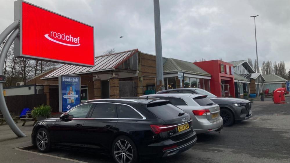 Single-storey service station building in brown, grey and red. There is a red Roadchef sign on a display screen to the left. Cars are parked outside the service station.  There is a red bin in the background.