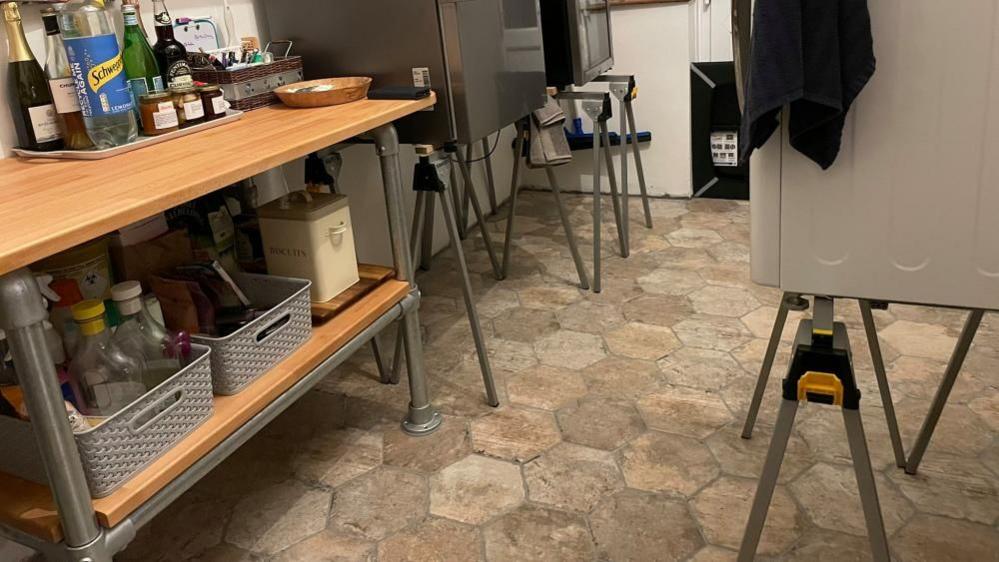 A photo of a kitchen where metal trestles are holding up a fridge in the background and a washing machine in the foreground. On the left of the image there is a kitchen side with jars and bottles. 