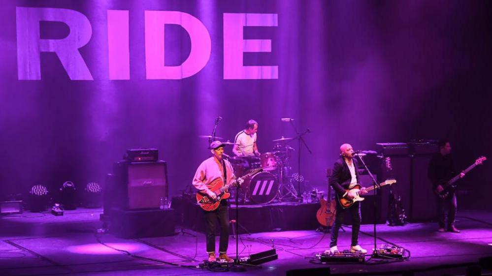 Ride perform at Barbican Hall, London. They are bathed in pink lighting, the band name emblazoned on the background of the stage.