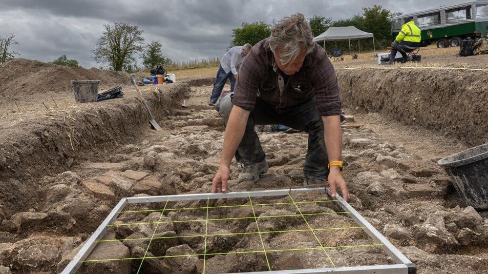 A man in an archaeology trench crouched on what looks like an old wall holding a square frame that appears to be for measuring.