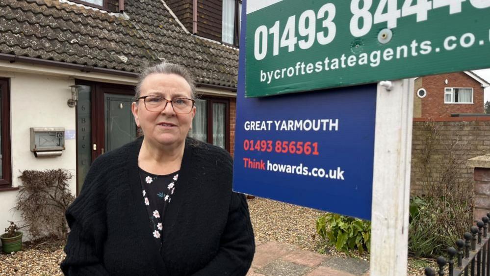 Marilyn Robinson standing in the garden of her home. Beside her are two estate agents boards indicating the property is for sale. She is standing in a garden where paving and gravel is laid. She is wearing a black cardigan and a black top with white flowers printed into the design. She is wearing glasses and her greying hair is tied back.