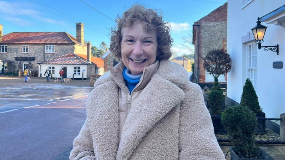 Lorraine Geary standing in the middle of a village, with short curly hair, she is smiling to the camera, wearing a blue top, with a cream jacket over the top and a cream fake fur coat. People and houses are behind her. 