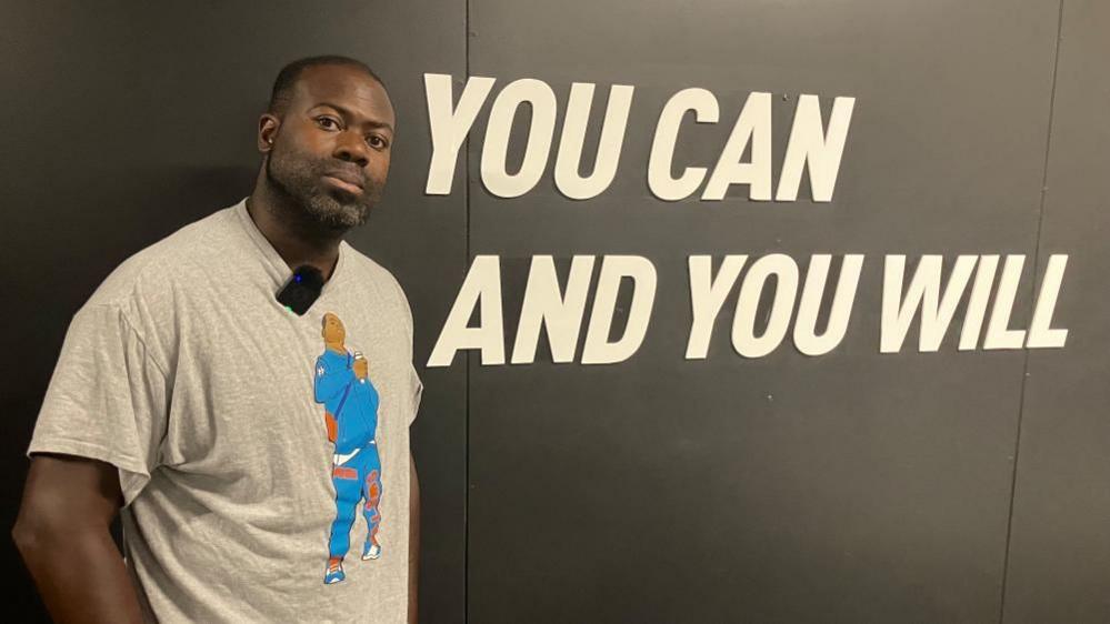 Swifty wearing a T-shirt in front of a sign that read "you can and you will"