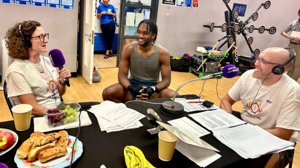 BBC Radio Bristol's Joe Sims and Claire Cavanagh (sat on the right and left respectively) sit around a table at Empire Fighting Chance, broadcasting the station's breakfast show. Joe smiles at Claire as she talks, as does a boxer sat between them. On the table sits various pieces of paper and plates of food.
