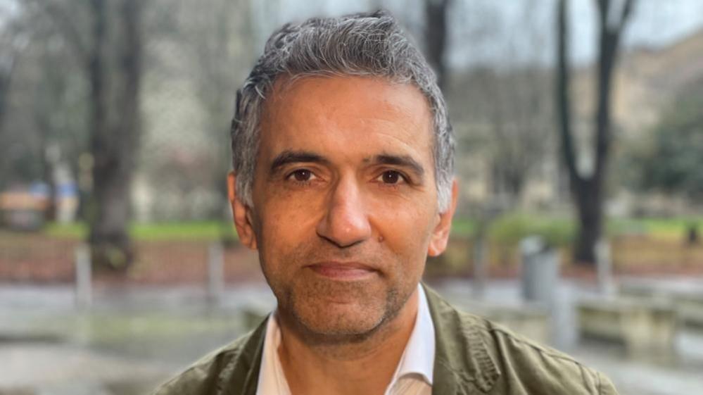 Prof Gurch Randhawa looks directly at the camera. He has grey hair and wears a white shirt and olive green jacket.