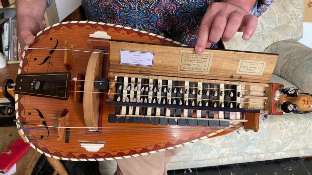 A hurdy-gurdy is being held while a man lifts open the instruments inner crank. It appears to be made of wood, with ornate details decorating its exterior. It is a mixture of red and brown in colour with lighter, yellow strings.