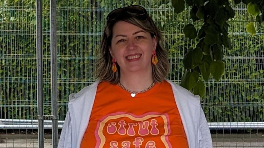 A woman stands in front of a temporary fence smiling at the camera. She is wearing an orange t-shirt that reads 'strut safe'