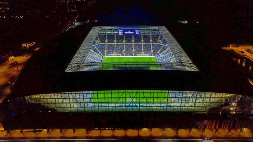 A drone view of Everton's new stadium, showing the pitch lit up at night.