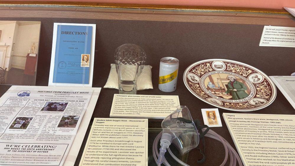 Inside a glass display case, with a can of Schweppes and a pamphlet of writing by Priestley