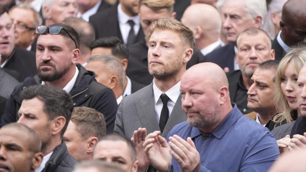 Andrew "Freddie" Flintoof stands among a large crowd of people. He wears a grey suit and has a solemn expression on his face.