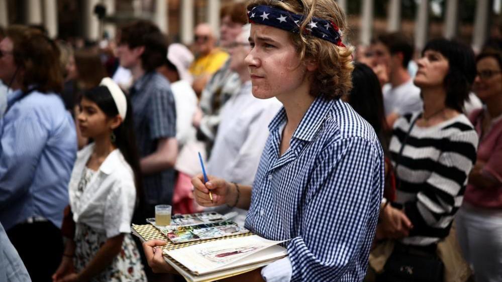 A young man wearing a headband stands in the crowd holding a paintbrush as he paints a picture of the Pope