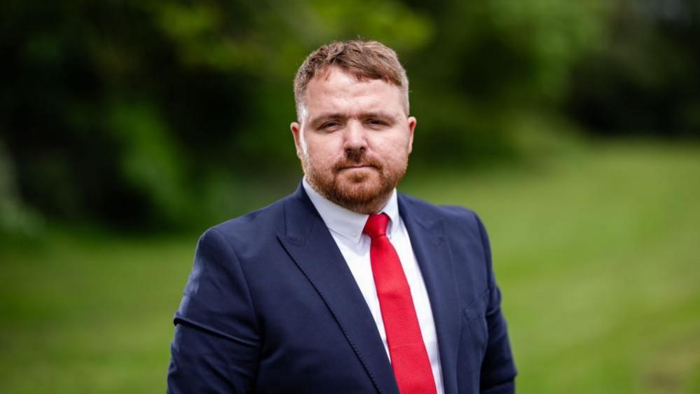 Robert Martin in a blue suit and red tie looking at the camera. He has light brown hair and beard.