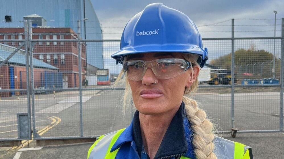 Donna Dryburgh wears a blue hard hat, which says babcock on it, and protective glasses. She appears to be standing in an industrial estate. She has a silver hoop nose piercing and her blonde hair is in a long plait. She wears blue overalls with a high vis vest over them.