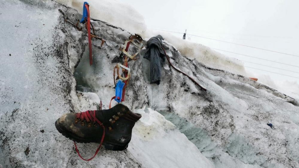 A brown, dirty boot with red shoelaces that belonged to a German climber who disappeared while hiking along Switzerland's Theodul Glacier in 1986. The boot and some other equipment like ropes are sitting in dirty snow.