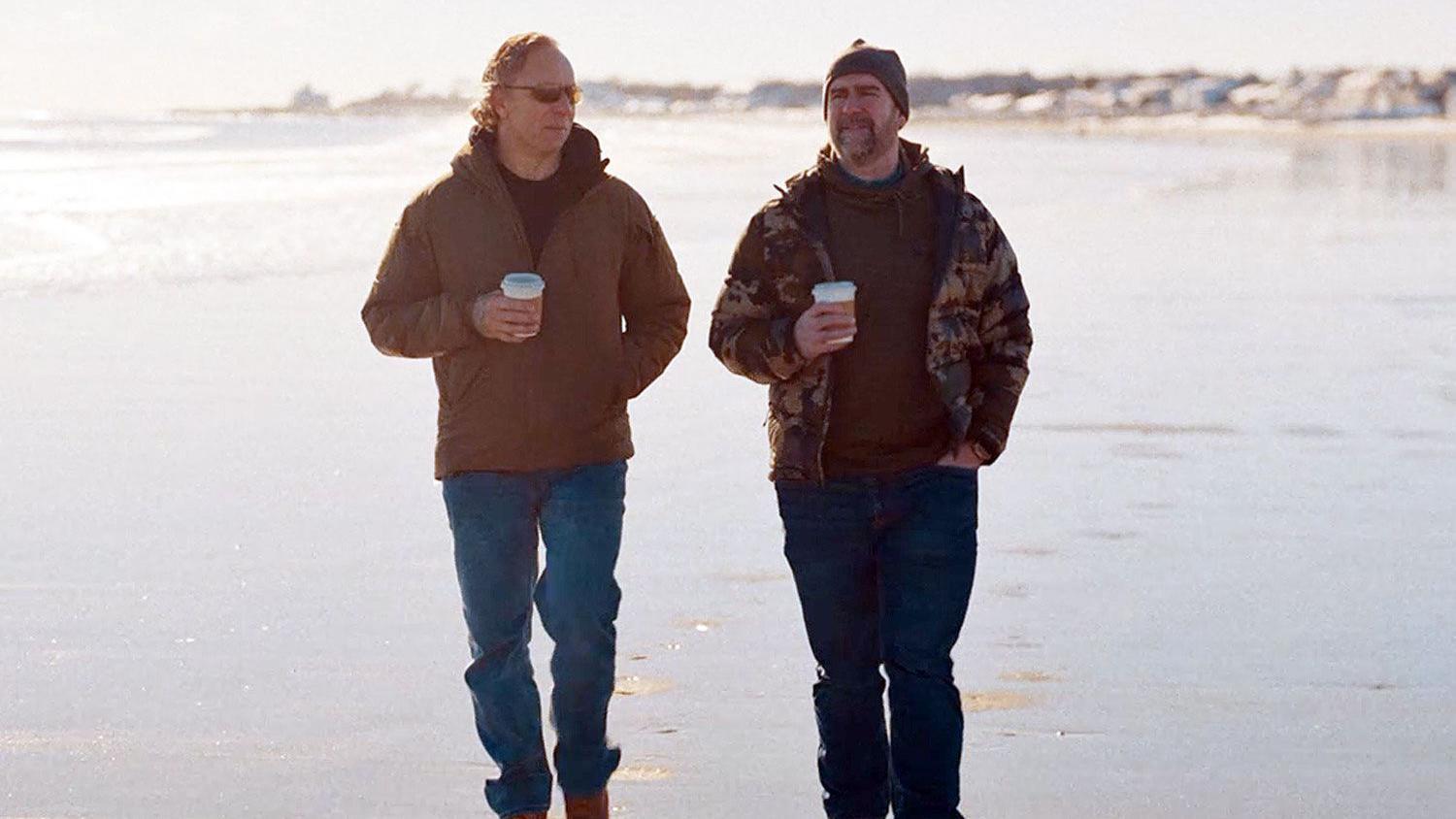 dvojica muškaraca šetaju plažom, u rukama im je kafa. Obojica nose plave pantalone i smeđe jakne.