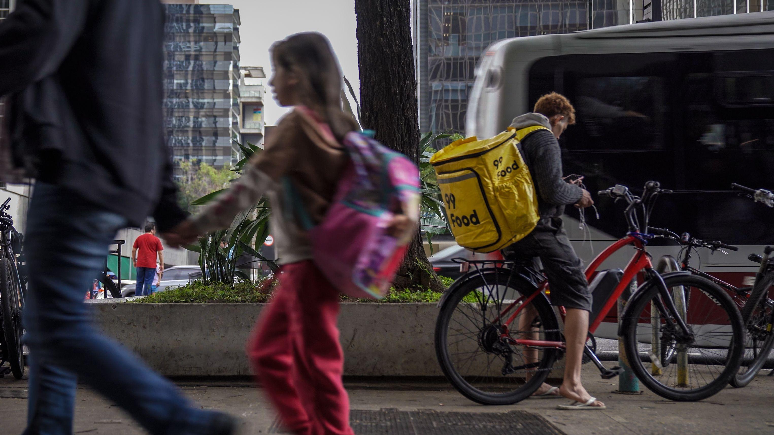 Homem e uma menina passando em movimento no primeiro plano; em segundo plano, com nitidez, um entregador est&aacute; em uma bicicleta vermelha, usando uma bag amarela da 99, olhando para o celular