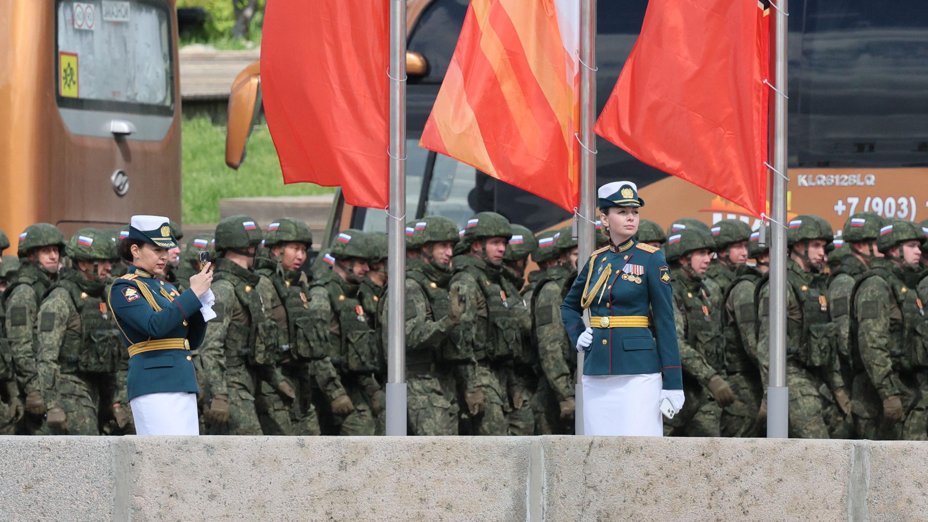 Dos militares rusas femeninas, vestidas con túnicas verdes adornadas con medallas, toman fotos mientras soldados con uniformes de combate verdes pasan el 7 de mayo de 2025. Banderas rojas y naranjas ondean en mástiles sobre el grupo.