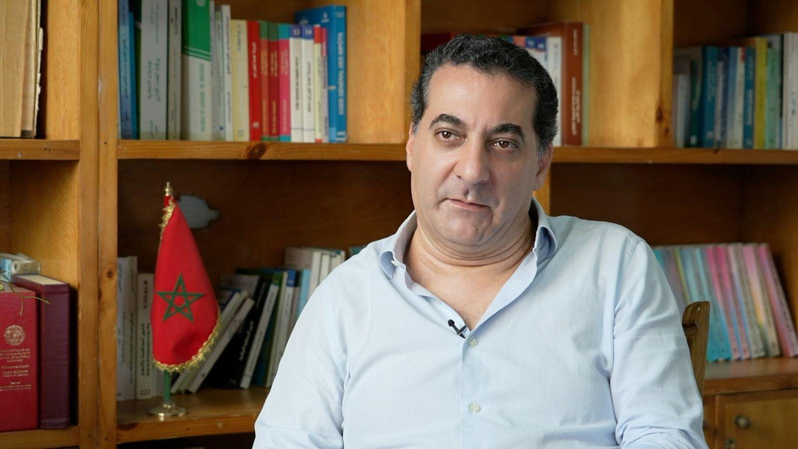 رجل جالس وخلفه مكتبة كتب وعلم المغرب