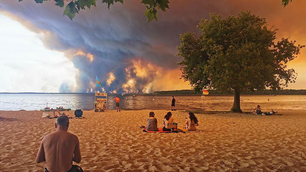 Turistas descansan en una playa francesa mientras en el horizonte se ve una nube de humo provocada por los incendios forestales. 