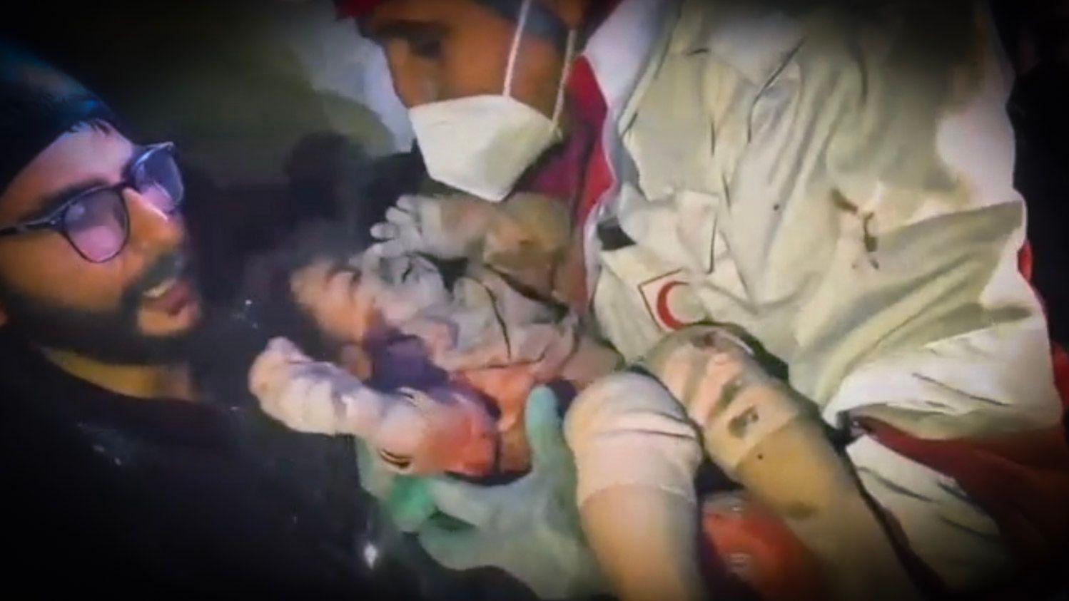 Um socorrista com m&aacute;scara segura uma crian&ccedil;a pequena coberta de poeira, enquanto outro observa