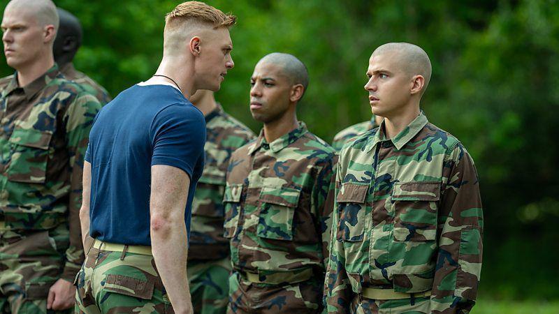 Diversos soldados de uniforme enfileirados com um bosque ao fundo, com o que parece ser um superior reclamando com eles