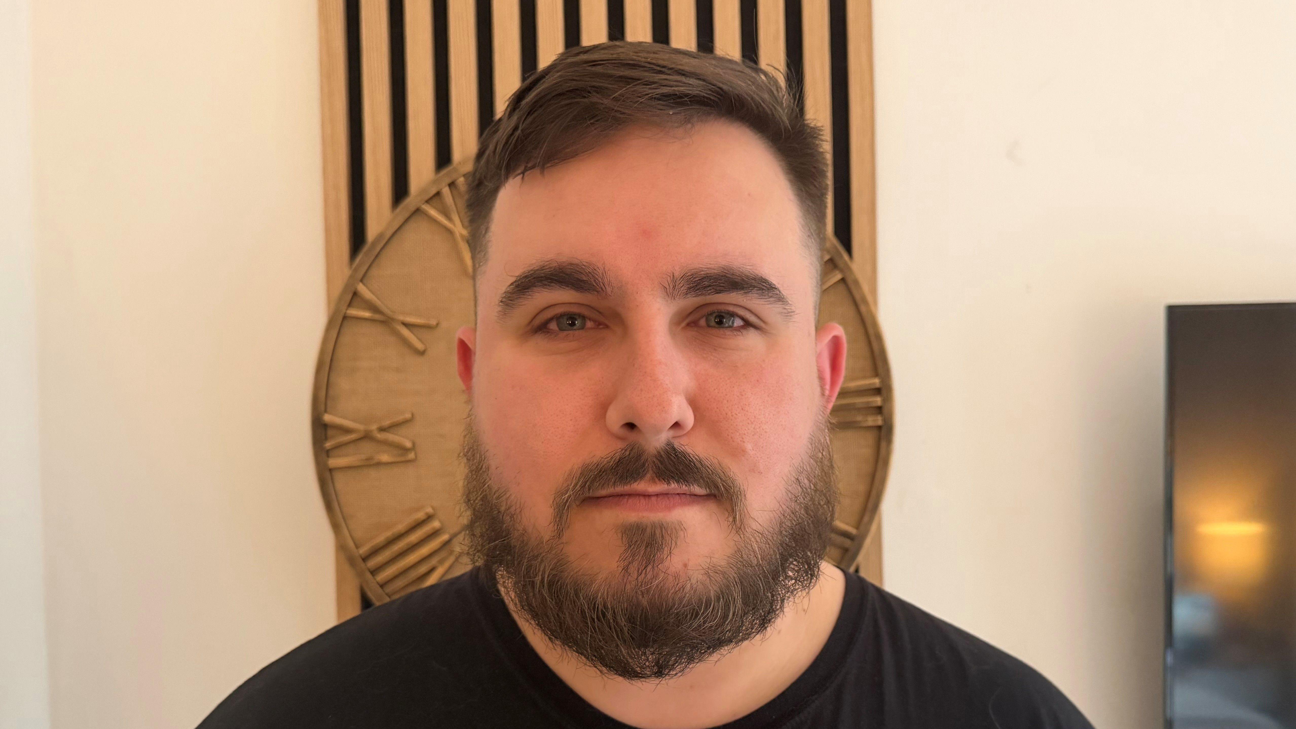 Primer plano de un joven, de medio cuerpo, con barba y bigote tupidos, vistiendo una camiseta negra. Tiene una expresión seria y hay un reloj de madera justo detrás de su cabeza.