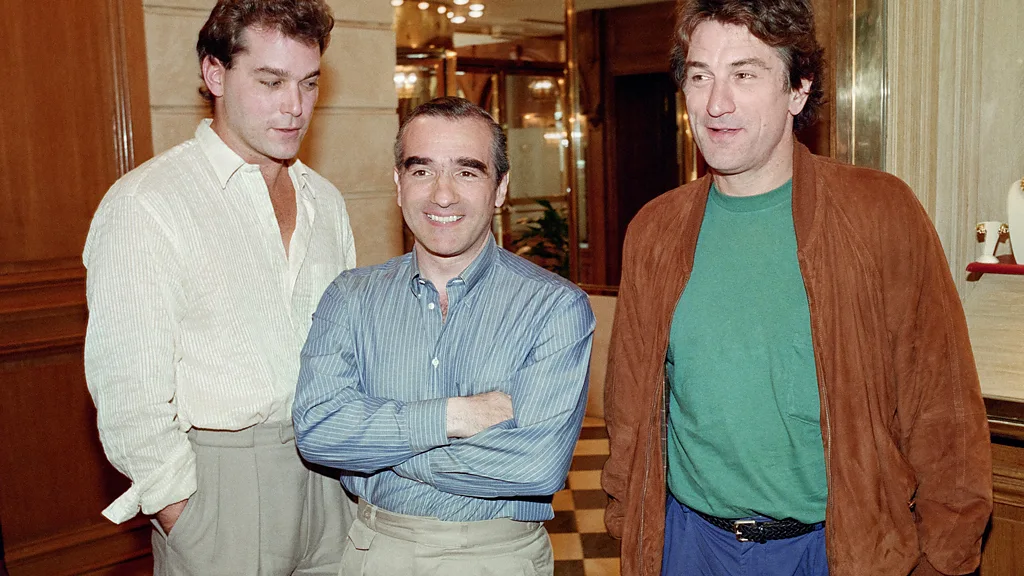 S leva na desno: Rej Liota u beloj košulji i svetlo sivim pantalonama gleda u Martina Skorsezea u sredini, koji je nasmejan u plavoj košulji na pruge, prekrštenih ruku i Robert De Niro, u zelenoj majici, smeđoj kožnoj jakni i plavim pantalonama gleda u daljinu