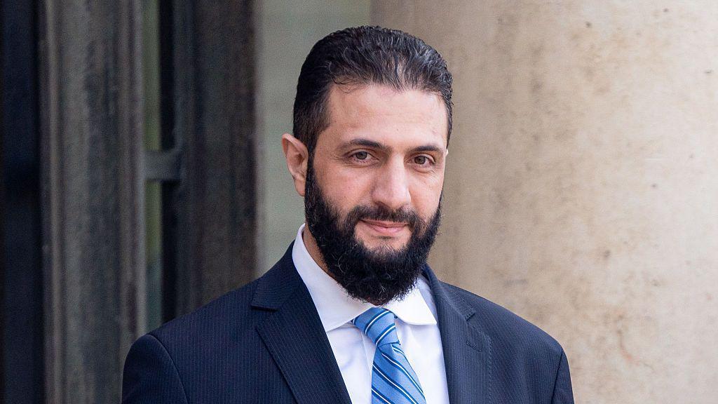 الشرع بلحية سوداء، مرتدياً بدلة رسمية كحلية اللون وقميص بلون أبيض وربطة عنق زرقاء مخططة، في قصر الإليزيه، يظهر خلفه حائط بلون سكري.