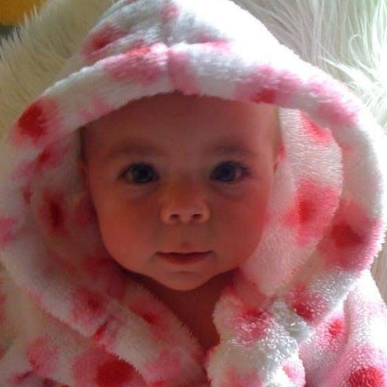 Sophia de bebé. Sonríe levemente a la cámara y lleva puesta una bata esponjosa, rosa y blanca, con la capucha puesta.