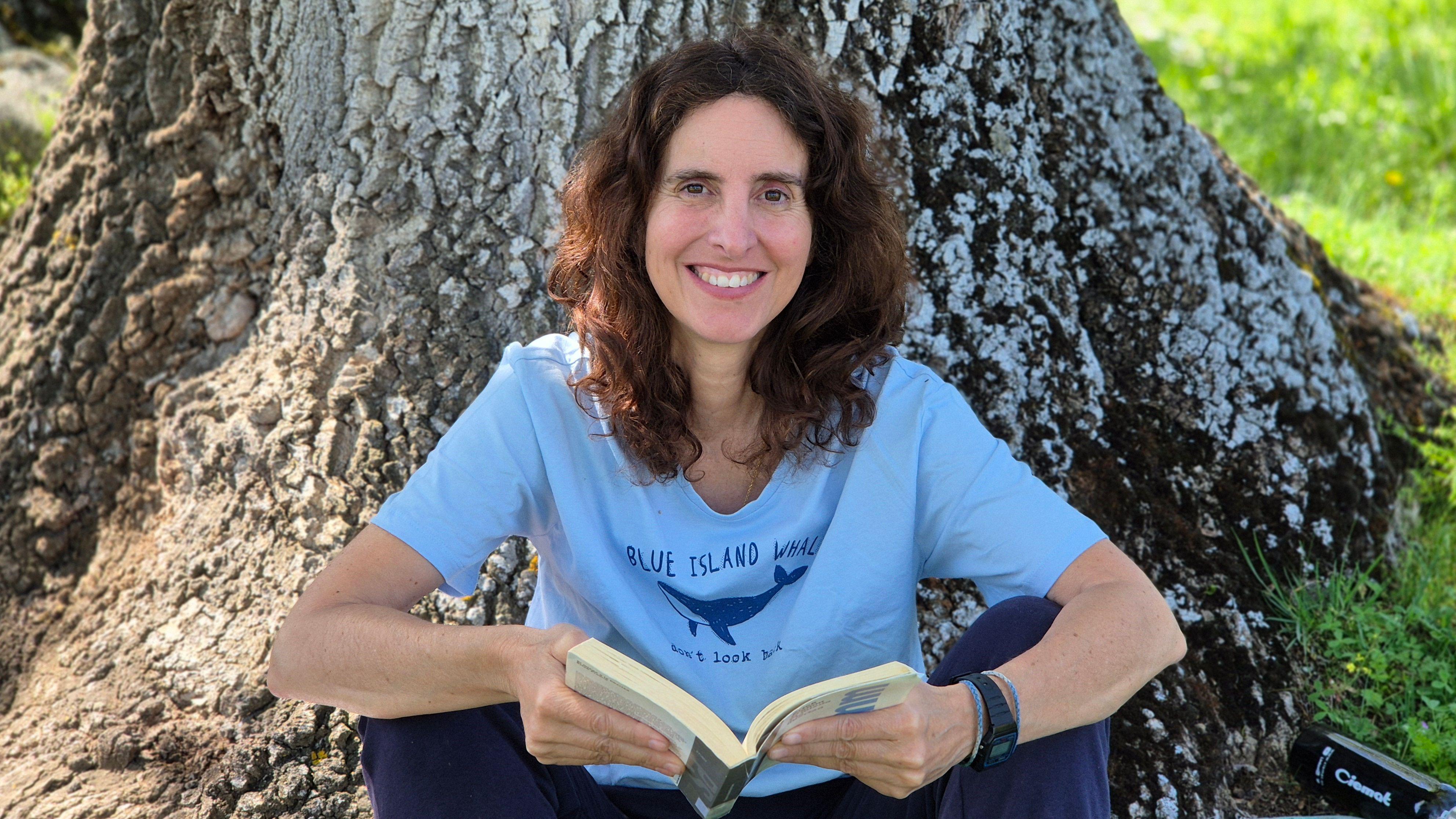 Laura G. De Rivera con un libro en la mano sonriendo con una camiseta azul enfrente de un árbol