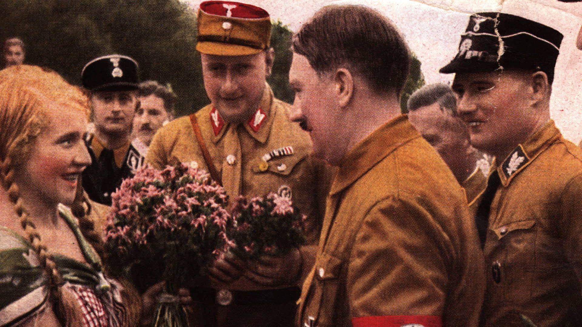 Uma mulher de cabelos loiros tran&ccedil;ados olha com admira&ccedil;&atilde;o para Hitler enquanto ele fala com ela, cercado por homens em uniformes militares. 