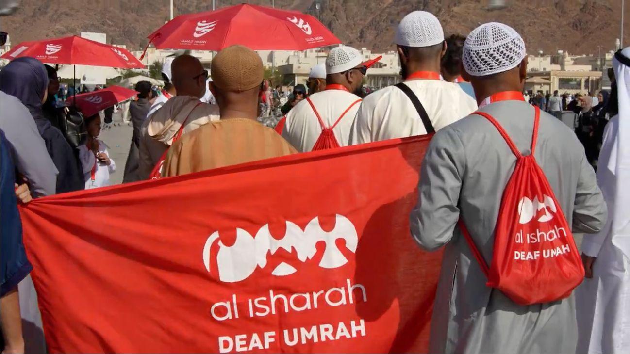 مجموعة من الرجال يرتدون زياً إسلامياً، يظهرون بظهورهم إلى الكاميرا، يحملون علماً أحمر كُتب عليه "al isharah DEAF UMRAH" باللون الأبيض مع رموز للغة الإشارة. ويحمل أحدهم حقيبة حمراء على ظهره تحمل العبارات والرموز نفسها.