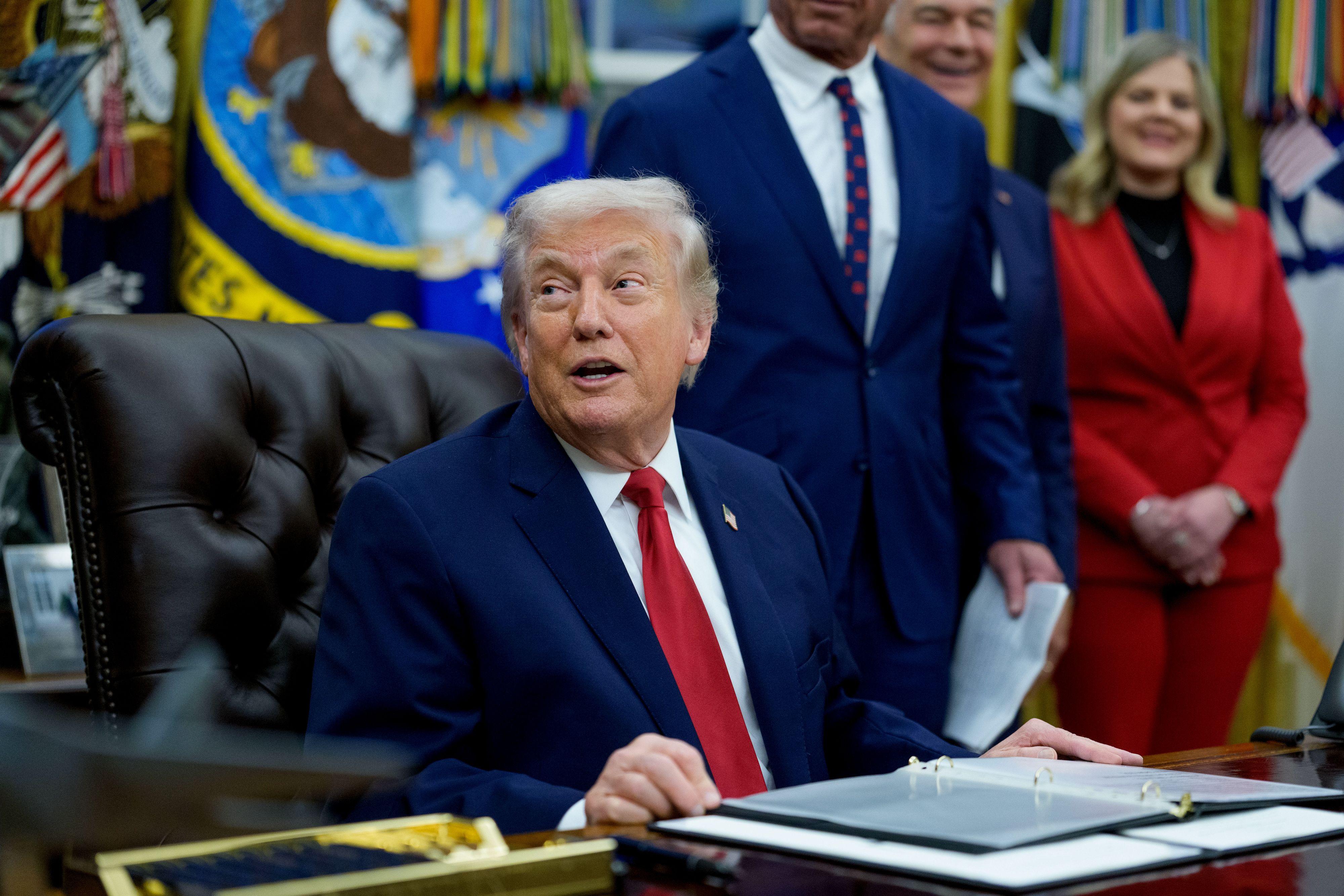 El presidente Donald Trump mira hacia un costado mientras está sentado en el escritorio del Despacho Oval rodeado de personas