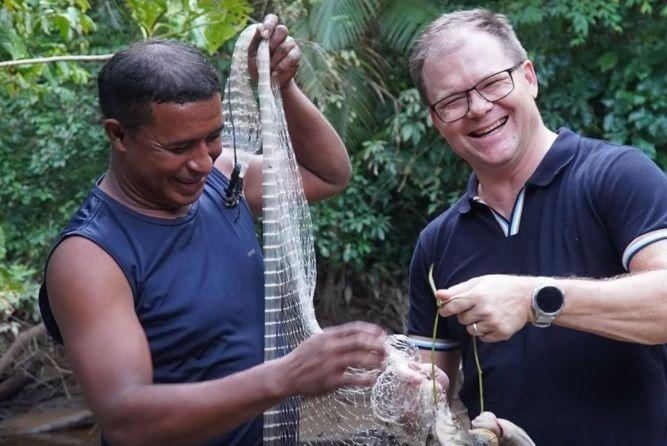 Dois homens com rede de pescador na floresta. Um segura rede e outro segura peixes.