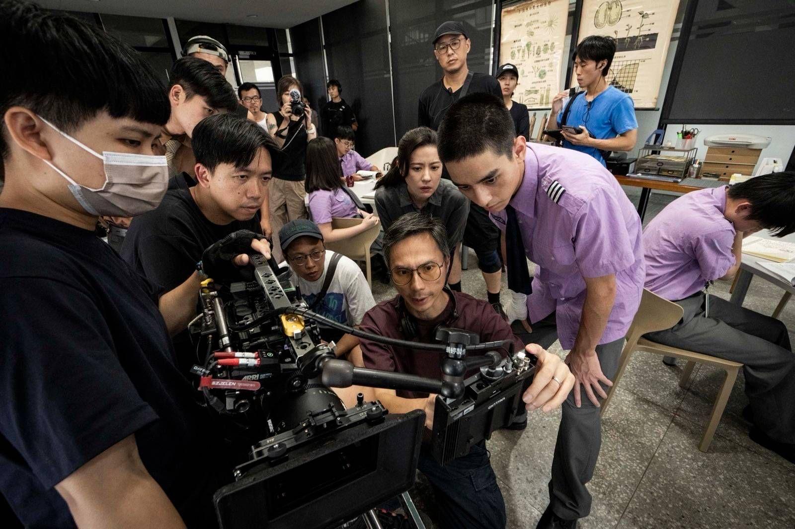 周冠威身穿棗紅色上衣身處片場中，他與演員及工作人員盯着攝錄機。