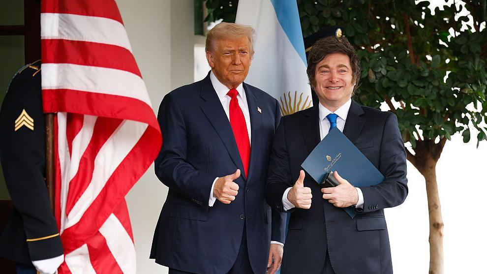 Donald Trump y Javier Milei, ambos de traje, saludan con sus pulgares arriba y las banderas de sus países en la Casa Blanca