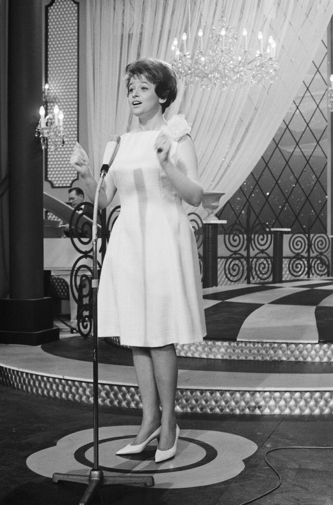 Una fotografía en blanco y negro de Marion Rung en el escenario frente a un micrófono de pie. Lleva un vestido ligero hasta la rodilla y zapatos de tacón de color claro. Detrás, hay un escenario con dos escalones, suelo ajedrezado, una cortina y una lámpara de araña de cristal.